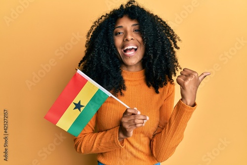 African american woman with afro hair holding ghana flag pointing thumb up to the side smiling happy with open mouth
