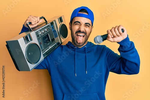 Young hispanic man holding boombox, listening to music singing with microphone sticking tongue out happy with funny expression.