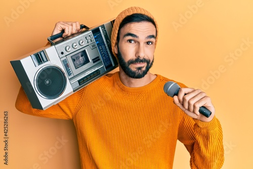 Young hispanic man holding boombox, listening to music singing with microphone relaxed with serious expression on face. simple and natural looking at the camera.