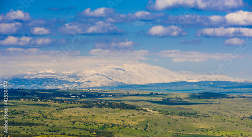 Beautiful winter landscape of Golan Heights: view of snow-capped Mount Hermon on a border with Syria and Lebanon - Israel's only ski resort, with green fields and clouds