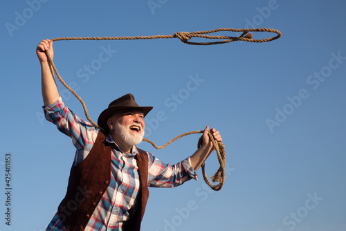 Old cowboy with lasso rope at ranch or rodeo. Bearded western man with brown jacket and hat catching horse or cow.