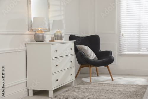 White chest of drawers, armchair and mirror in room. Interior design