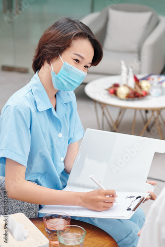 Young Asian nurse in medical mask collecting anamnesis when talking to patient in clinic lounge area