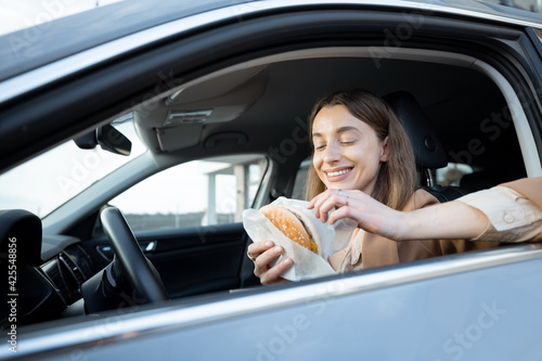Happy woman eating a burger in the car. Have unhealthy fast food snack. Food to go. Hungry and busy concept.