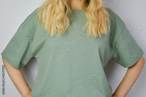 Blond hair woman posing near a light wall. Beautiful young caucasian girl. Hand gestures. Emotion. Casual clothing. Studio model in work. Strong woman, future is female. Jeans and green blank t-shirt