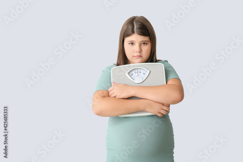 Sad overweight girl with measuring scales on light background