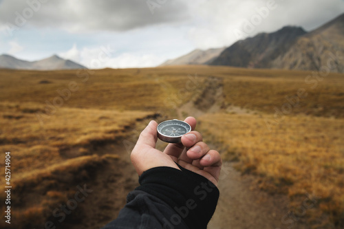 Man's hand holding a magnetic compass first-person view against the background of a high-altitude path and mountains