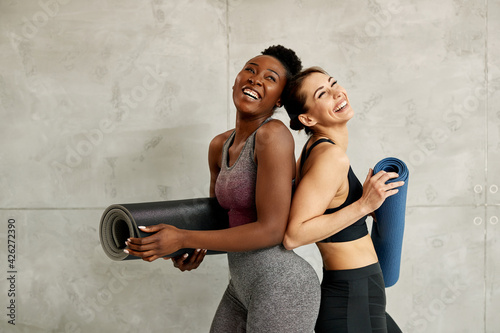 Young cheerful athletic women holding Yoga mats and laughing against the wall.