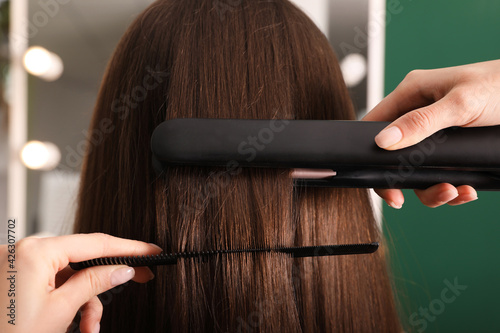 Stylist straightening woman's hair with flat iron in salon