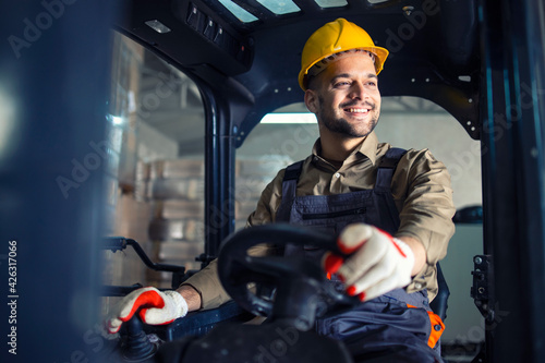 Young caucasian male in working uniform and yellow hardhat operating forklift machine in warehouse storage room.