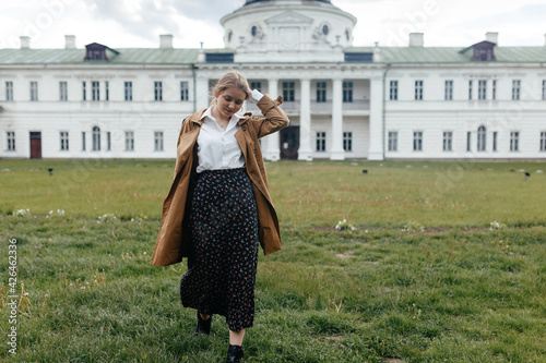 Stylish young girl walks on the grass against the background of a white old manor house and looks down
