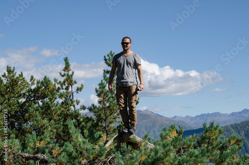A young man stands on the top of a tree against a background of mountains