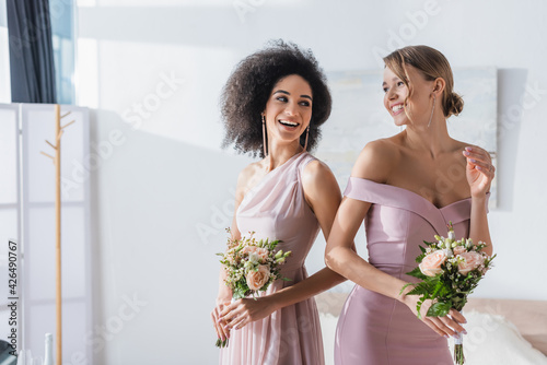 joyful multicultural bridesmaids holding wedding bouquets in bedroom.