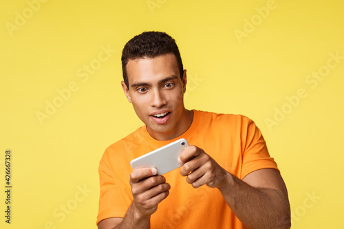 Excited handsome young male student waste time playing cool new game, holding smartphone horizontally, turn body as trying win rase, smiling joyful, download awesome app, standing yellow background
