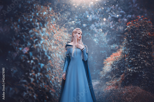 Beautiful fantasy woman elven goddess walks in spring nature forest. Long creative vintage blue dress, sleeves. Blond hair fluttering in motion. Fairy cute face. Art Girl elf princess fashion model.