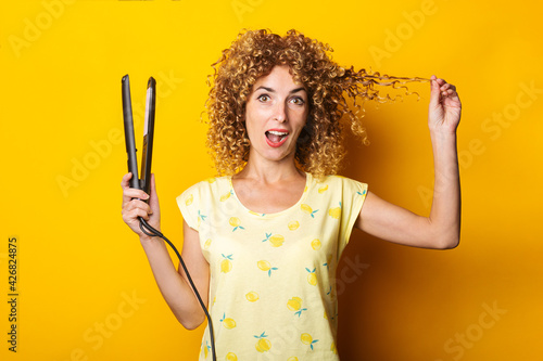 surprised young woman with curly hair with hair straightener on yellow background