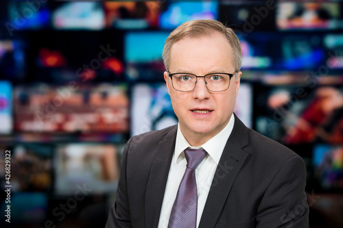 An anchorman reporting live breaking news sitting in Tv studio. Background of multiple screens of broadcast control room. Journalism concept