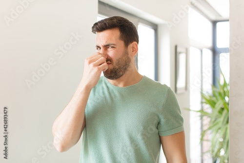 handsome man feeling disgusted, holding nose to avoid smelling a foul and unpleasant stench