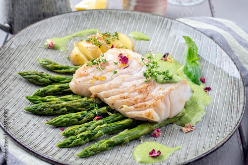 Modern style traditional fried skrei cod fish steak with green asparagus and potatoes served as close-up on a Nordic style plate