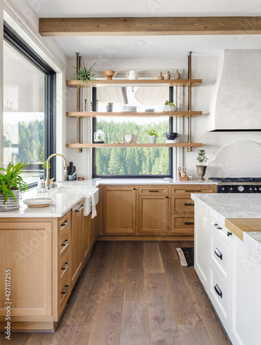 Traditional kitchen in new luxury home with farmhouse sink, hardwood floors, wood beams, large island and quartz counters. Features large windows inviting abundant natural light, and floating shelves.