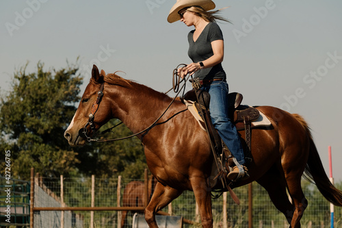 Western cowgirl lifestyle shows woman in cowboy hat riding mare horse through outdoor arena during summer in Texas.