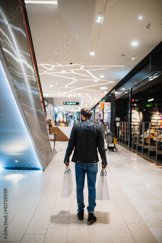 Back view of a young caucasian man in a black leather jacket with bags in his hand walks in the mall during the epidemic COVID - 19 coronavirus