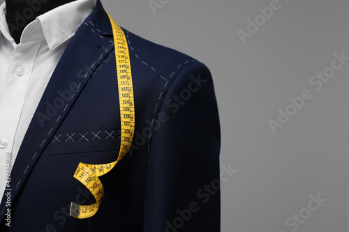 Semi-ready jacket with tailor's measuring tape on mannequin against grey background, closeup. Space for text