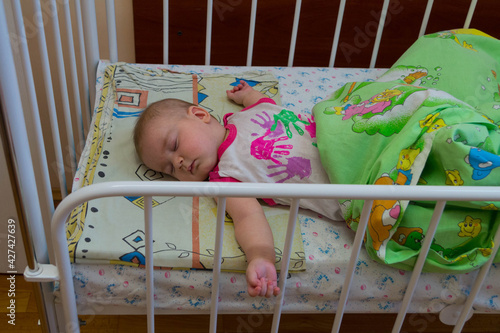 A small child sleeps, covered with a blanket, in a white metal bed in a hospital or orphanage