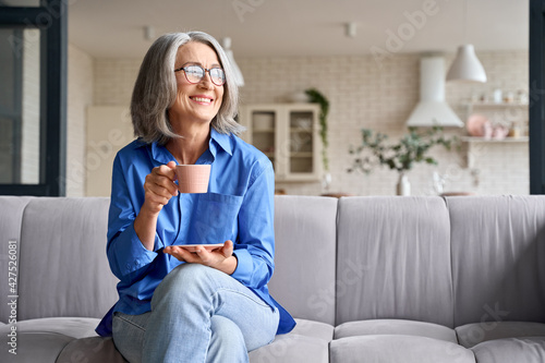 Smiling mature elder 60s woman sitting relaxing with cup of tea, coffee. Senior mid age stylish look woman with eyeglasses portrait with cup looking away at modern home.