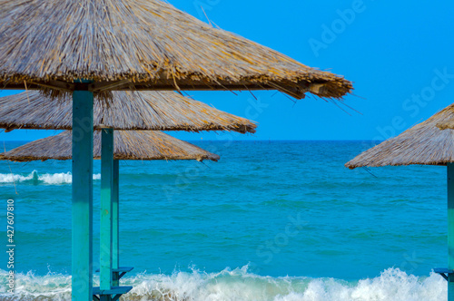 Close-up of straw beach umbrellas providing shade near the wavy water of the Black Sea on sandy shoreline on a sunny clear summer day at Vama Veche, Romania