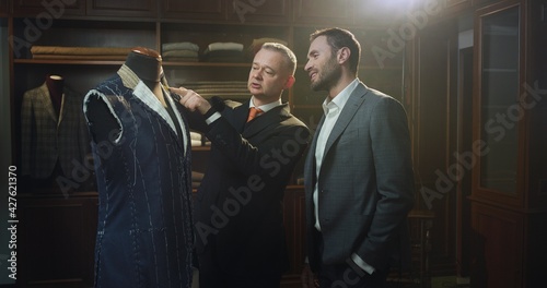 Cinematic shot of young handsome man is trying his new custom tailored suit with designer in a luxury tailoring atelier. Concept of fashion, handmade, hand craft, couturier and business.