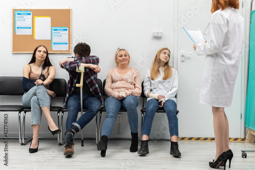 The doctor standing in front of her office reads out the next person and invites them in for a check-up. People of different age are sitting on chairs