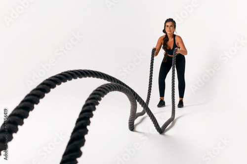 Sporty young woman in black suit doing crossfit exercises with ropes over isolated white background. hard workout. wave