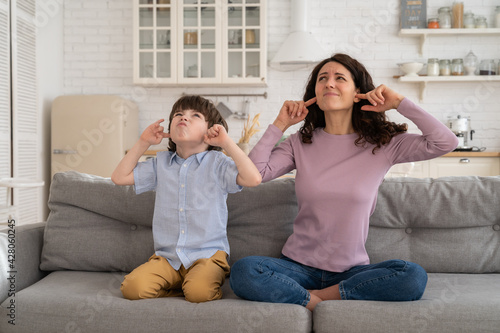 Frowning mom and son sit on couch with closed eyes and cover ears from noisy music or fight sounds from neighbors. Young female parent and kid tired of drilling noise not listen plug ears with fingers