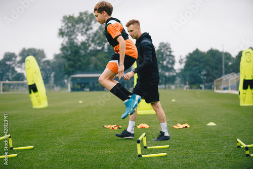 Young man jumping over practice hurdles in soccer training. Soccer coach watching boy on training. Player of youth football academy with coach on pitch