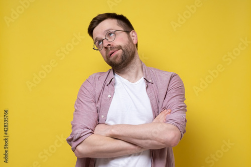 Caucasian man in glasses and casual clothes looks up distantly with arms crossed, he is bored and uninteresting. Yellow background.