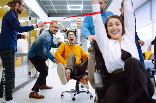 Friendly work team ride chairs in office room cheerfully excited diverse employees laugh while enjoying fun work break activities, creative friendly workers play a game together.