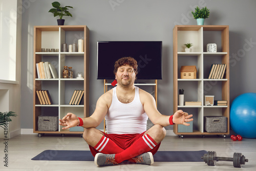Overweight fat millennial man in sportswear with eyes closed relaxing on lotus position meditating calm down practicing yoga sitting on mat at living room. Home exercise, sports activity on quarantine