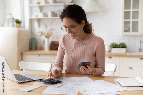 Young latina female work with financial papers at home count on calculator before paying taxes receipts online by phone. Millennial woman planning budget glad to find chance for economy saving money