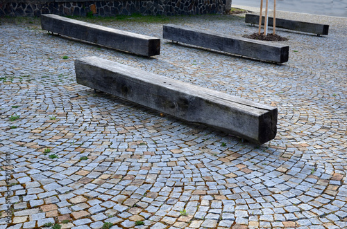 square with park benches made of natural oak wooden prisms. trunk cut into a block shape. granite paving and stone walls. trees in decorative circles