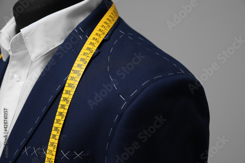 Semi-ready jacket with tailor's measuring tape on mannequin against grey background, closeup