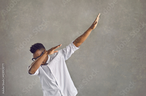 Happy handsome young black man with Afro hairstyle dancing dub against gray stone street wall. Funky African American guy in white shirt doing popular dab dance arm move on concrete urban background