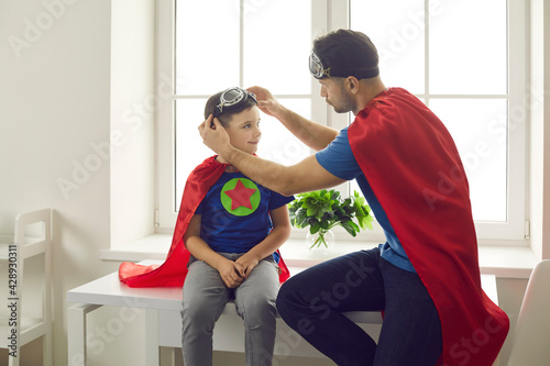 Happy family dressed in super hero costumes having fun at home. Father and child wearing red superhero capes spending time together. Dad helping son put on aviator goggles while playing games at home