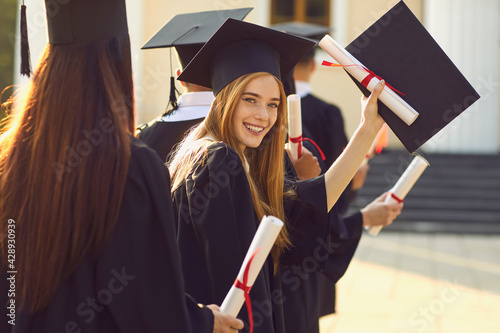 Successful graduation from university. Smiling beautiful girl university or college graduate standing with diploma and looking at camera over mates around and university at background