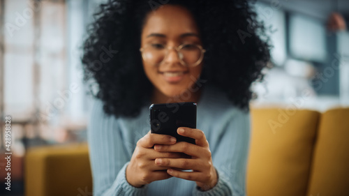 Portrait of a Beautiful Authentic Latina Female with Afro Hair in Stylish Cozy Living Room Using Smartphone at Home. She's Browsing the Internet and Having Fun. Focus on Mobile Phone.