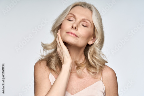 Gorgeous senior older woman with closed eyes touching her perfect skin. Beautiful portrait mid 50s aged woman advertising facial antiage lift products salon care tighten skin isolated on white.