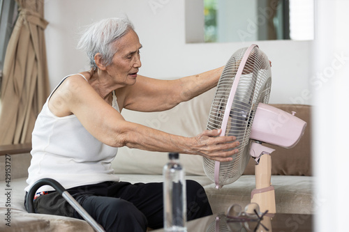 Overheated asian senior woman sweating,high temperature in sunny day while stay at home,cooling herself in front of an electric fan,old elderly suffering from heat,hot summer weather,lifestyle concept