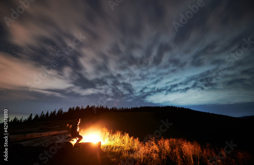 Side view of human relaxing near cozy bonfire and looking at starry sky. Concept of resting on nature with wonderful view blue night sky. Concept of travelling, hiking and night camping.