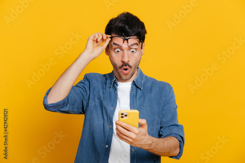 Amazed shocked caucasian guy holding smartphone in his hand, looking at the phone in surprise with his glasses raised, stunned facial expression, stands on isolated orange background