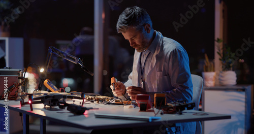 Professional adult caucasian repairman doing technical repairment circuit board soldering for broken drone device in dark office laboratory interior. Engineering,